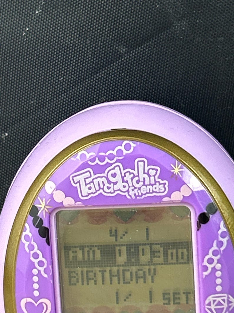 May include: A purple Tamagotchi Friends digital pet toy with a gold trim. The screen displays the time and the word "BIRTHDAY". The device has decorative elements including hearts and jewels.