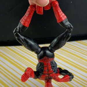K&ouml;nnte beinhalten: Eine rot-schwarze Spider-Man-Actionfigur mit detailliertem Kost&uuml;mdesign. Die Figur wird kopf&uuml;ber gehalten und zeigt ihre beweglichen Gliedma&szlig;en und die ikonische Maske. Die roten Bereiche haben ein Netzmuster.