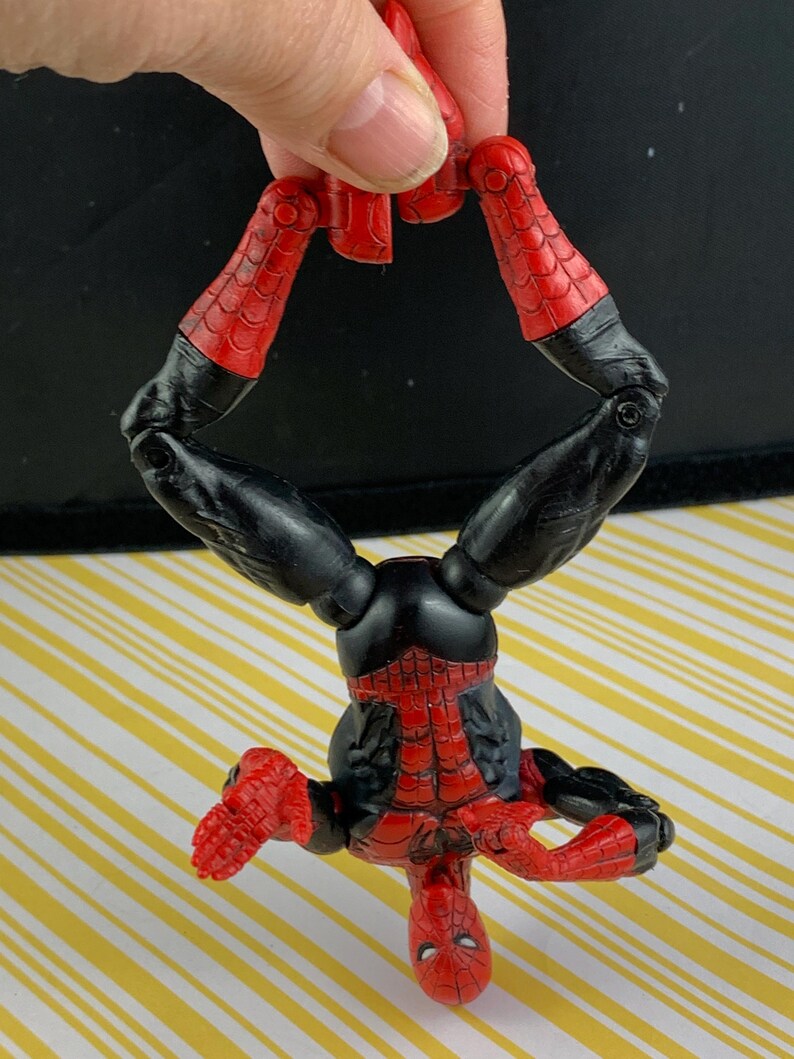 K&ouml;nnte beinhalten: Eine rot-schwarze Spider-Man-Actionfigur mit detailliertem Kost&uuml;mdesign. Die Figur wird kopf&uuml;ber gehalten und zeigt ihre beweglichen Gliedma&szlig;en und die ikonische Maske. Die roten Bereiche haben ein Netzmuster.