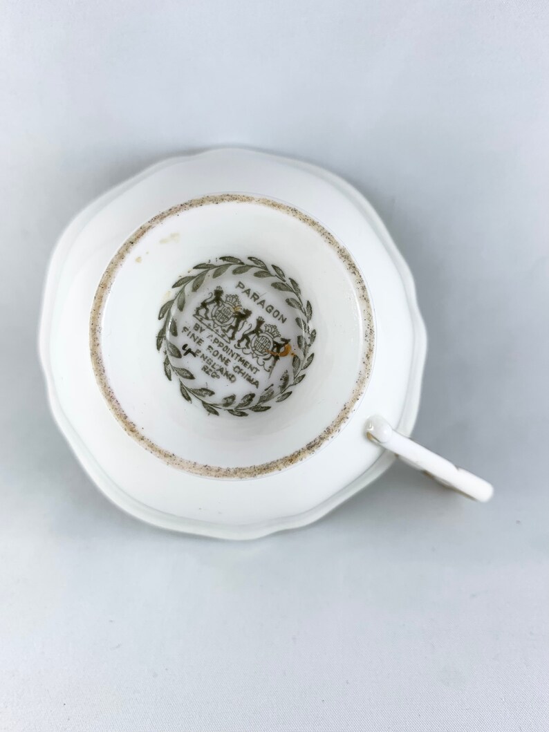K&ouml;nnte beinhalten: Eine wei&szlig;e Teetasse mit einem gr&uuml;n-schwarzen Logo, auf dem "Paragon by Appointment to Her Majesty Fine Bone China England" steht, auf der Unterseite.
