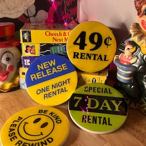 May include: A collection of five round, colorful buttons with different rental prices and terms. The buttons are yellow, blue, green, and white with black text. The text on the buttons reads "49¢ Rental", "New Release One Night Rental", "Special 7 Day Rental", and "Please Rewind Be Kind".