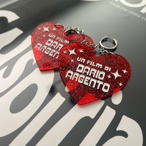 May include: Two red glitter heart-shaped keychains with silver key rings. Each keychain features white text that reads "Un Film Di Dario Argento" with a star design.