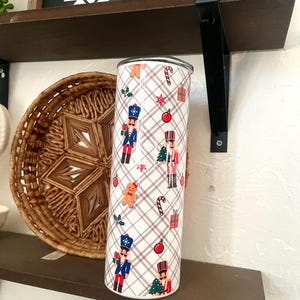 May include: A white, stainless steel tumbler with a festive Christmas theme. The tumbler features a repeating pattern of nutcrackers, Christmas trees, ornaments, and gifts on a plaid background. The tumbler is approximately 8 inches tall.