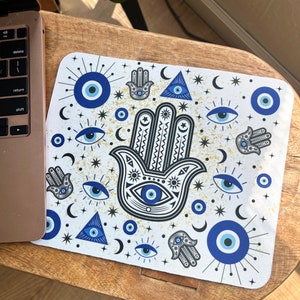 May include: A white mouse pad with a blue and black hamsa hand design. The hamsa hand is surrounded by blue evil eyes, crescent moons, and stars. The background is a light gold color.