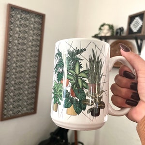 May include: White ceramic mug with a geometric design featuring a variety of potted plants in shades of green. The mug has a black handle.