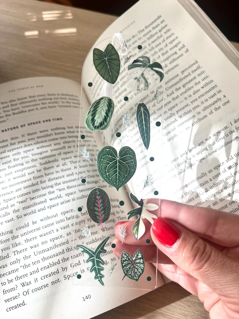 Pu&ograve; includere: Un segnalibro trasparente con una variet&agrave; di illustrazioni di piante verdi e bianche. Il segnalibro &egrave; progettato per assomigliare a una finestra con le piante che sembrano essere dietro il vetro.