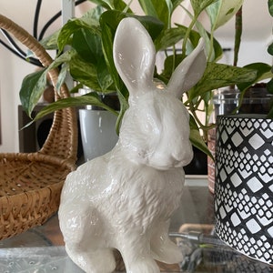 May include: A white ceramic rabbit figurine with a textured fur design. The rabbit is sitting with its ears upright and its head tilted down.