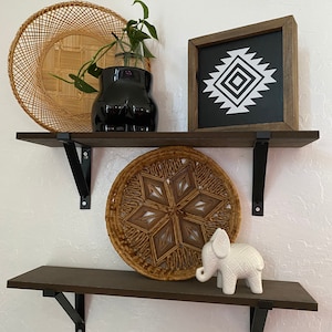May include: Two dark brown shelves with black brackets are mounted on a white wall. The top shelf holds a woven basket, a black vase with greenery, and a framed black and white geometric design. The bottom shelf displays a woven starburst and a white elephant figurine.