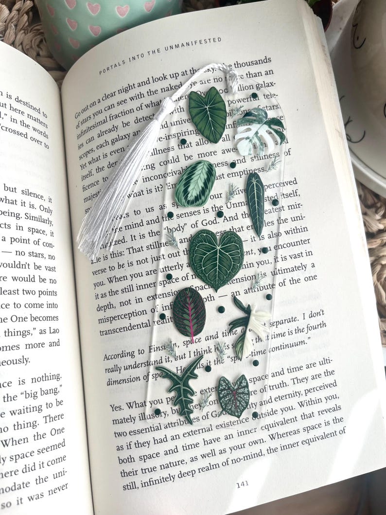 Pu&ograve; includere: Un segnalibro con nappa bianca con foglie e fiori pressati verdi e bianchi. Il segnalibro &egrave; su un libro aperto con testo sull'universo e lo spazio. Il testo dice "Esci in una notte limpida e guarda le migliaia di stelle che puoi vedere a occhio nudo. Non siamo altro che una frazione infinitesimale di ci&ograve; che si trova oltre noi."