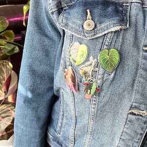 May include: A denim jacket with five different plant-themed pins. The pins are in the shape of hearts and leaves. The pins are all different colors and have a glossy finish.