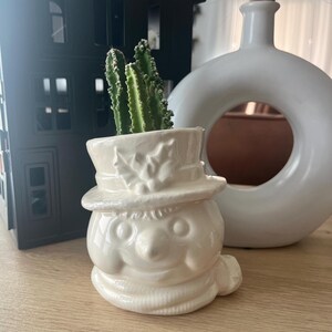 May include: White ceramic snowman planter with a green cactus plant inside. The snowman has a black top hat, a black nose, and black eyes. The planter is sitting on a wooden surface.