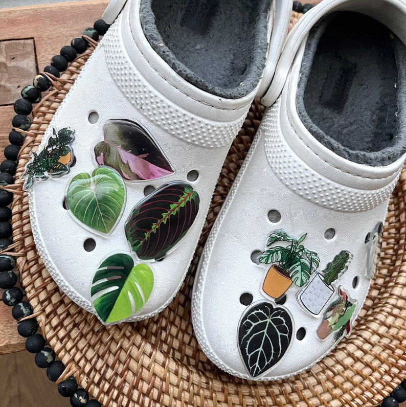 May include: White clogs with various green, pink, and black plant stickers. The stickers are of different shapes and sizes, including leaves, flowers, and potted plants.