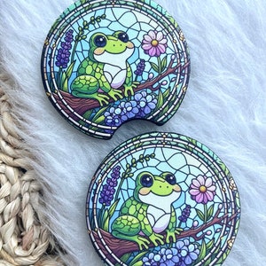May include: Two round green frog car coasters with a stained glass design. The frogs are sitting on branches with purple and pink flowers.