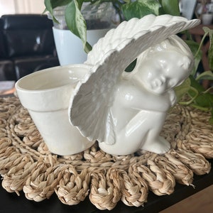 May include: White ceramic planter shaped like a sleeping angel with wings. The planter is sitting on a woven straw mat.