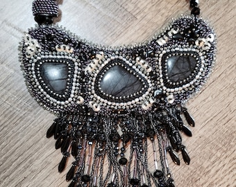 May include: A black beaded necklace with a crescent-shaped design and a fringe of black beads. The necklace features three large, dark grey stones.