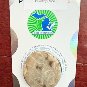 May include: A white card with a green and blue Michigan state outline sticker. The card says "Petoskey Stone" and "Made in Michigan". The card also features a round, tan-colored phone grip with a brown swirl pattern. The phone grip says "PopGrip" in pink, blue, and black letters. The card also has a holographic sticker with the text "Swappable PopTop Interchangeable".