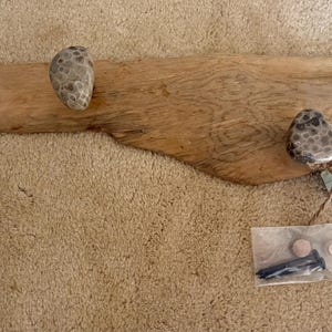 May include: A rustic wooden coat rack with two stone knobs and two wooden pegs. The driftwood-style rack has a natural wood grain and is accompanied by a small tag and hardware.