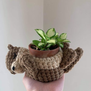 May include: A handmade crochet squirrel planter in shades of brown and tan, holding a small terracotta pot with a green and white plant. The squirrel has a detailed face and is lying on its back.