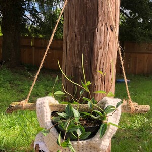 May include: A white crocheted sloth planter hanging from a wooden post with a green plant growing inside. The planter is suspended by twine.