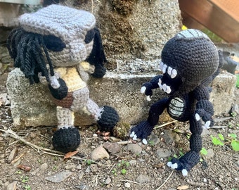 May include: Two handmade crochet figures of characters from the Alien and Predator movie franchises. The Predator is gray, tan, and brown, while the Xenomorph is black with white accents. Both are posed on a concrete surface.