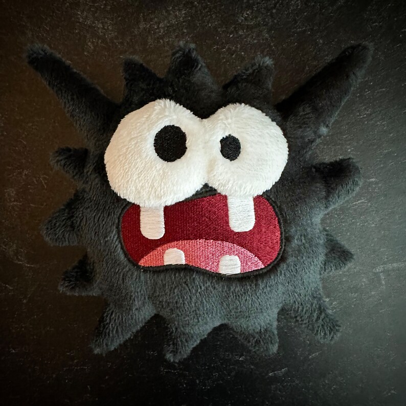 Peut inclure: Un jouet en peluche noir avec une texture h&eacute;riss&eacute;e et duveteuse. Il a une grande bouche ouverte avec des dents blanches et une langue rouge. Le jouet a de grands yeux blancs avec des pupilles noires.