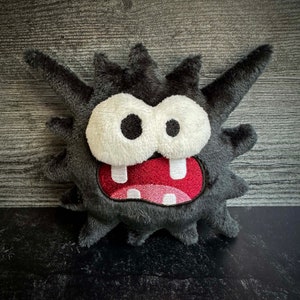 Peut inclure: Un jouet en peluche noir avec une texture h&eacute;riss&eacute;e et poilue. Il a une grande bouche ouverte avec des dents blanches et une langue rouge. Les yeux sont grands et blancs avec des pupilles noires.