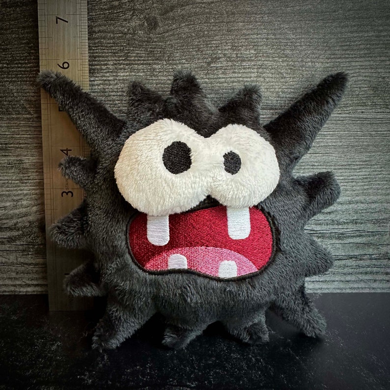 Peut inclure: Un jouet en peluche noir et gris en forme de monstre avec un grand sourire aux dents et des yeux blancs. Le monstre a une texture h&eacute;riss&eacute;e et poilue.
