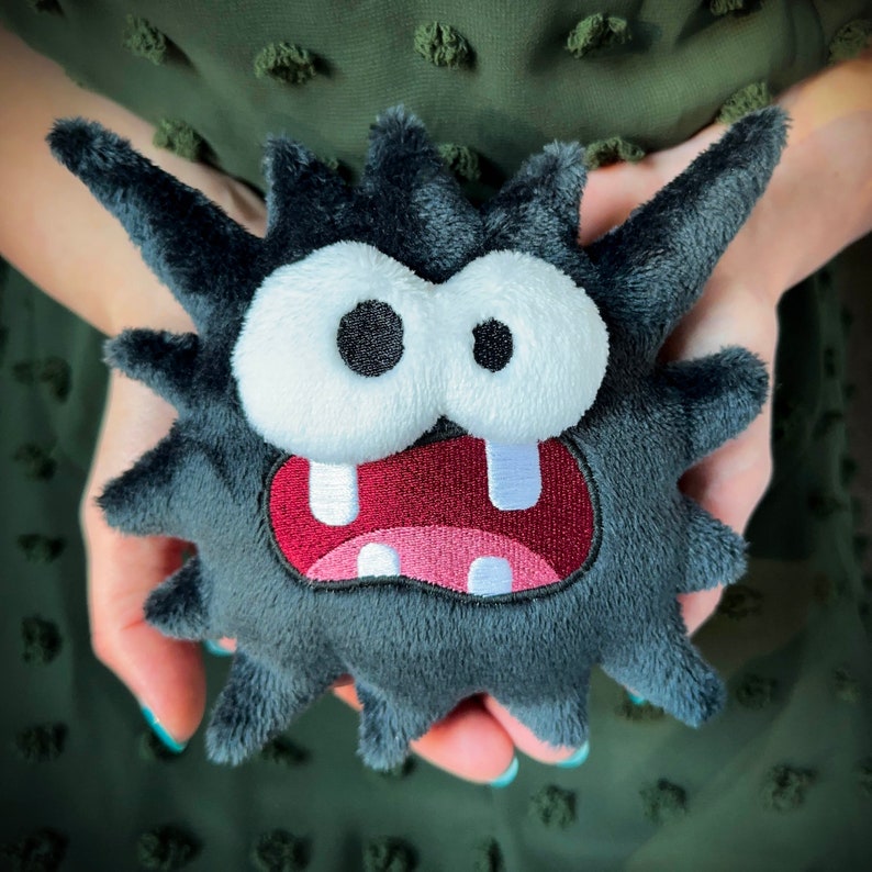 Peut inclure: Une cr&eacute;ature en peluche noire et grise avec un sourire narquois et de grands yeux blancs. La cr&eacute;ature a une bouche rouge et une langue rose.
