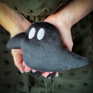 May include: A plush black crow with white eyes. The crow is held in the hands of a person wearing a green dress.