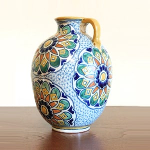 May include: A ceramic vase with a round body and a handle. The vase is decorated with a blue, green, orange, and yellow floral pattern.