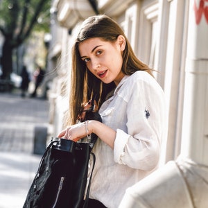 May include: A woman wearing a white button-down shirt and black pants is holding a black drawstring backpack. She is standing in front of a white building.