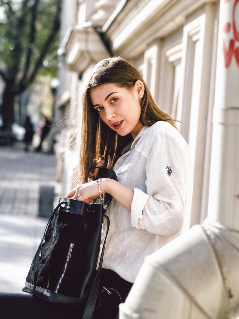 May include: A woman wearing a white button-down shirt and black pants is holding a black drawstring backpack. She is standing in front of a white building.
