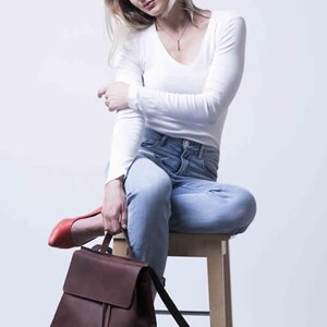 以下が含まれることがあります： 白い長袖シャツと青いジーンズを着用した女性が木製の椅子に座っています。彼女はフラップクロージャー付きの茶色の革のバックパックを持っています。バックパックは調節可能なストラップが付いています。