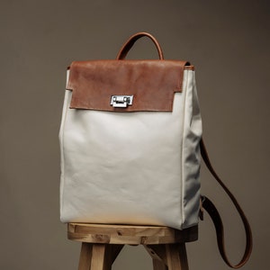 May include: A white canvas backpack with a brown leather flap and a silver buckle. The backpack is sitting on a wooden stool.