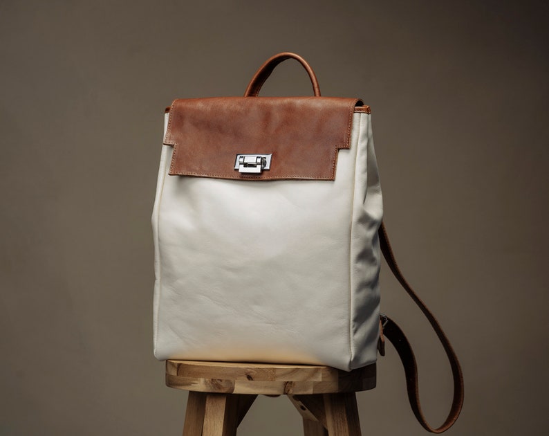 May include: A white canvas backpack with a brown leather flap and a silver buckle. The backpack is sitting on a wooden stool.