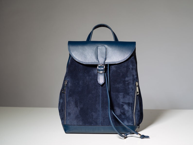 May include: A dark blue suede backpack with a leather flap and buckle closure. The backpack has two zippered pockets on the sides and a drawstring closure at the top.