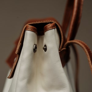 May include: Close-up of a white leather bag with brown leather straps and silver metal accents. The bag has a flap closure and a top handle.