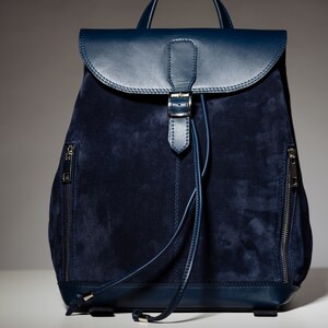 May include: A dark blue suede backpack with leather accents and a buckle closure. The backpack has two zippered pockets on the sides.