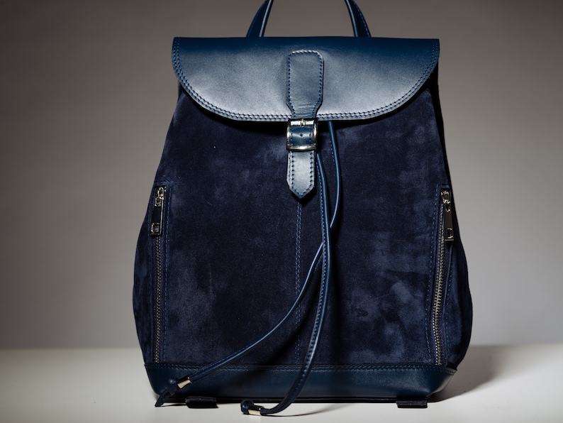 May include: A dark blue suede backpack with leather accents and a buckle closure. The backpack has two zippered pockets on the sides.