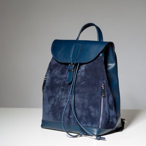 May include: A blue suede and leather backpack with a drawstring closure and a buckle on the front flap.