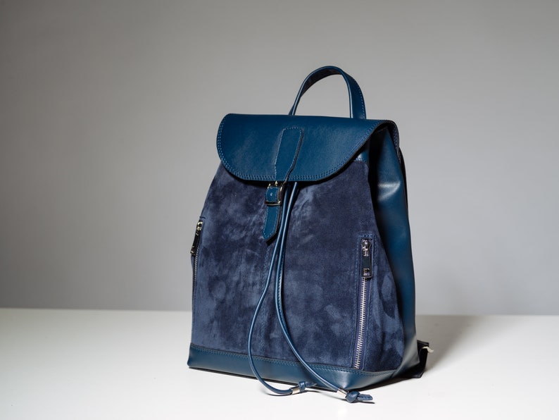 May include: A blue suede and leather backpack with a drawstring closure and a buckle on the front flap.