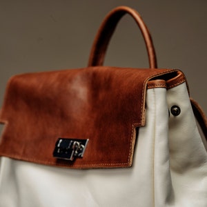 May include: A brown leather satchel with a white leather flap and a silver clasp. The bag has a white leather strap and a white leather handle.