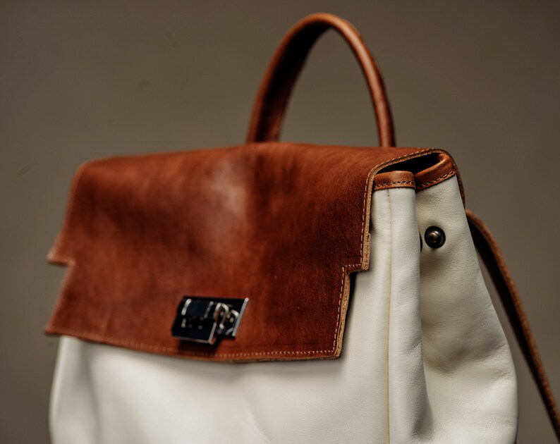 May include: A brown leather satchel with a white leather flap and a silver clasp. The bag has a white leather strap and a white leather handle.