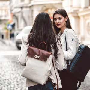May include: Two women walking down a cobblestone street. One woman is wearing a white jacket and a black backpack. The other woman is wearing a white jacket and a brown and white backpack.