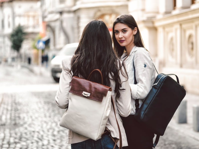 May include: Two women walking down a cobblestone street. One woman is wearing a white jacket and a black backpack. The other woman is wearing a white jacket and a brown and white backpack.