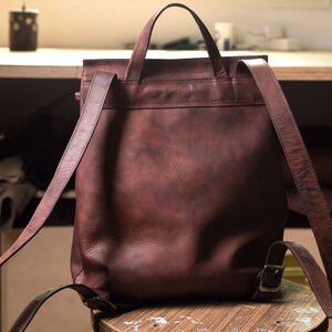 以下が含まれることがあります： 調節可能なストラップとバックル留め具が付いた茶色の革製バックパック。バックパックはフラップ留め具とトップハンドルが付いています。