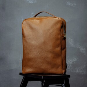 Könnte beinhalten: Ein brauner Lederrucksack mit Reißverschluss und goldenem Reißverschluss. Der Rucksack steht auf einem schwarzen Holzhocker.