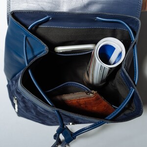 May include: A blue suede and leather backpack with a zippered pocket and a drawstring closure. The backpack is open and contains a rolled up magazine, a phone, and a brown leather wallet.
