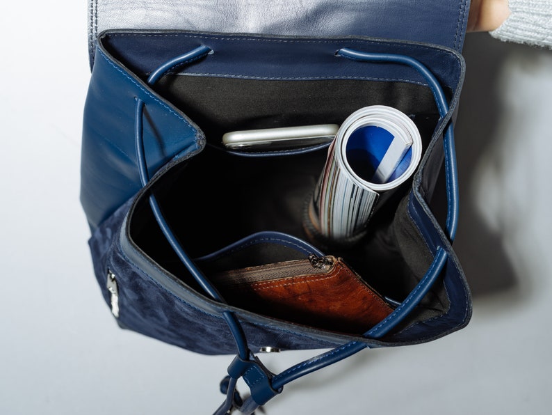 May include: A blue suede and leather backpack with a zippered pocket and a drawstring closure. The backpack is open and contains a rolled up magazine, a phone, and a brown leather wallet.