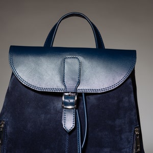 May include: A dark blue suede backpack with a leather flap and buckle closure. The backpack has a top handle and zippered pockets on the sides.